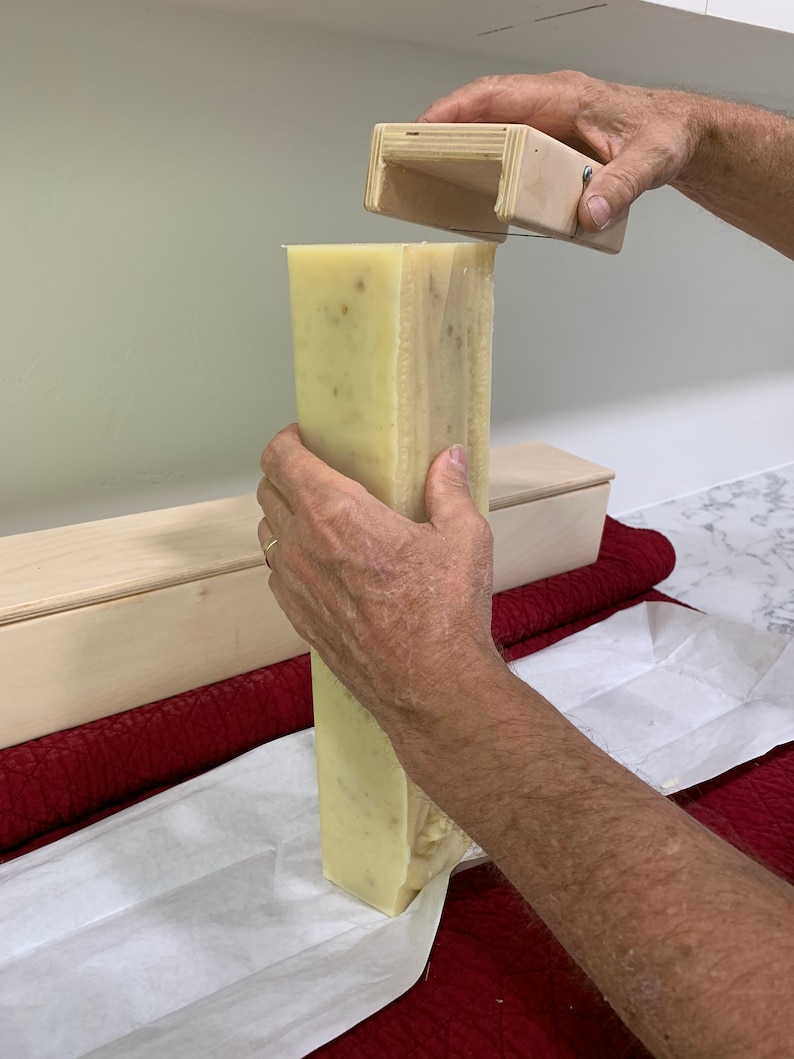 K&ouml;nnte beinhalten: Eine Person schneidet einen gro&szlig;en Block selbstgemachter Seife mit einem h&ouml;lzernen Seifenschneider. Die Seife ist hellgelb und hat eine k&ouml;rnige Textur. Die Seife liegt auf einem wei&szlig;en Blatt Papier.
