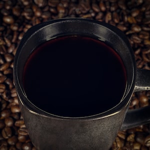 May include: A close-up of a black ceramic mug filled with dark coffee. The mug is sitting on a bed of roasted coffee beans.