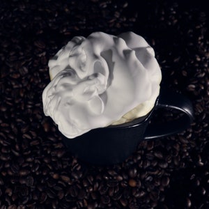 May include: A black coffee mug with a thick layer of white whipped cream on top. The mug is sitting on a bed of coffee beans.