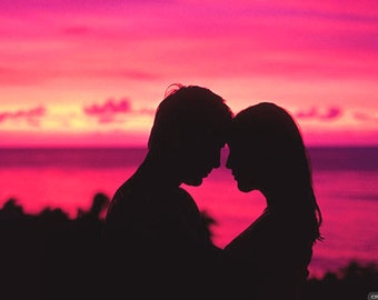 May include: Silhouette of a couple embracing against a vibrant pink and orange sunset. The couple is standing on a hill overlooking a body of water.