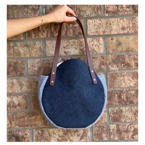 May include: A navy blue felt tote bag with brown leather handles. The bag has a rounded shape with a patterned fabric trim in shades of gray and white. The handles are attached with silver rivets.