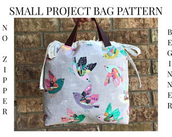 May include: A small project bag with a bird pattern. The bag is light gray with colorful birds in flight. It has brown leather handles and the text "SMALL PROJECT BAG PATTERN" and "NO ZIPPER BEGINNER" in black.