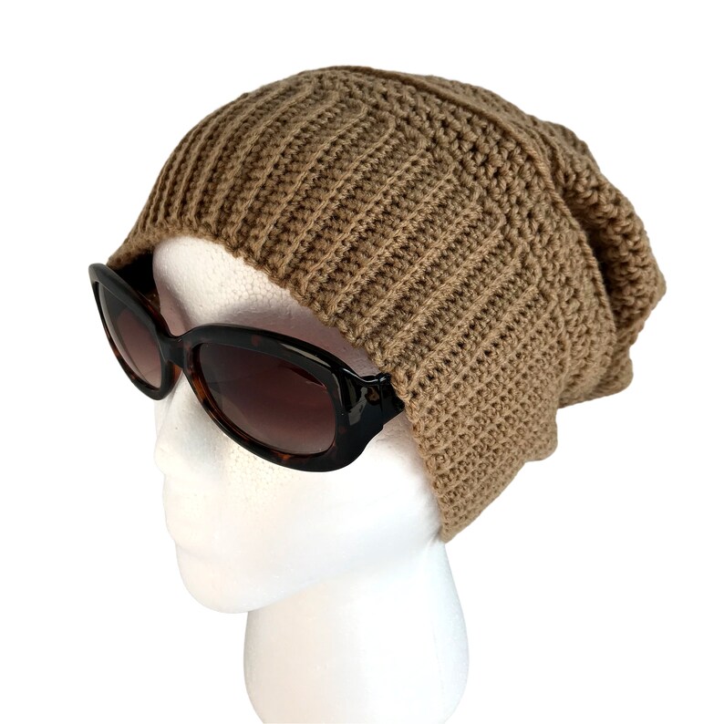May include: A brown crocheted beanie hat on a white mannequin head. The hat has a slouchy fit and is made with a simple stitch pattern.