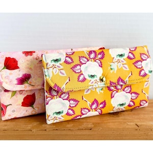 May include: Two fabric wallets with floral patterns. One wallet is pink with red flowers and the other is yellow with pink and white flowers. Both wallets have a button closure.