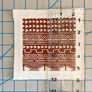 May include: A square fabric piece with a brown and white tribal pattern is measured with a clear ruler. The ruler shows measurements in inches. The fabric has a white border and is placed on a blue grid surface.