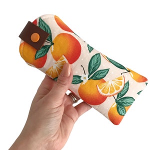 May include: A white fabric eyeglass case with a repeating pattern of orange slices and green leaves. The case has a brown leather strap with a small orange button.