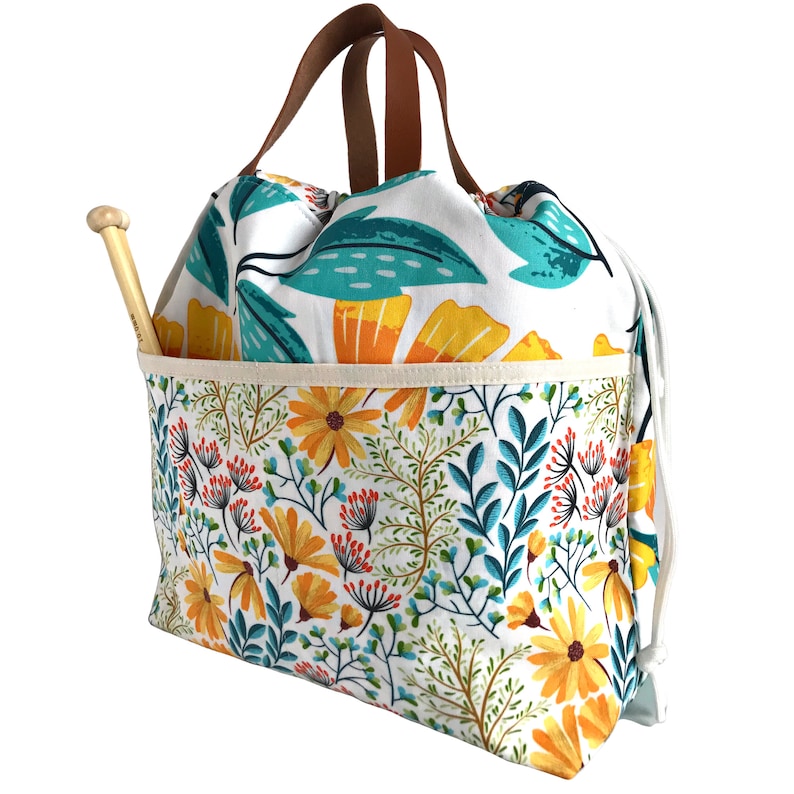 Puede incluir: Una bolsa de cord&oacute;n blanco con un patr&oacute;n floral en tonos de amarillo, naranja, turquesa y verde. La bolsa tiene un asa de cuero marr&oacute;n y un bolsillo separado en la parte delantera. Una aguja de tejer de madera sobresale del bolsillo.