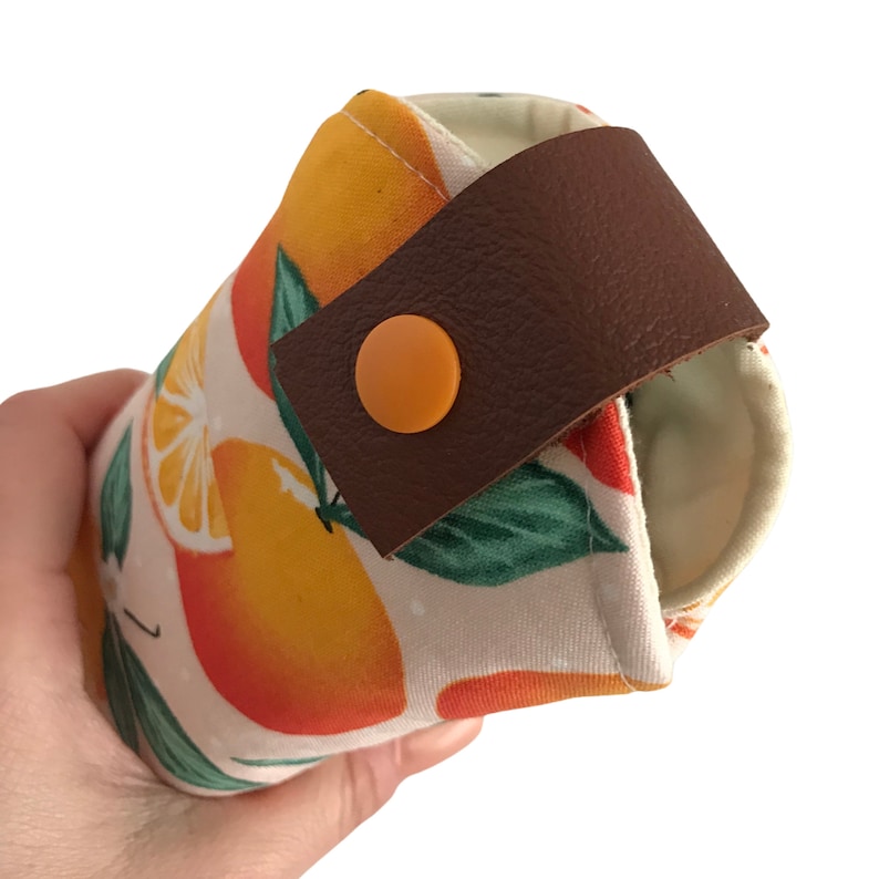 Pode incluir: Um porta-bebidas isolado de tecido branco com estampa de laranja, com uma al&ccedil;a de couro marrom e um fecho de bot&atilde;o laranja.