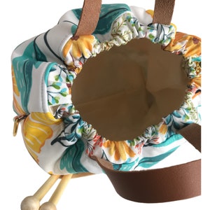 Puede incluir: Una bolsa de cord&oacute;n blanco con estampado floral en amarillo, naranja, rojo, azul y verde. La bolsa tiene asas de cuero marr&oacute;n y cierre de cremallera. Dos agujas de tejer de madera son visibles en la bolsa.