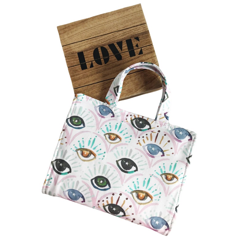 May include: A white tote bag with a colorful eye pattern. The bag has a brown wooden box with the word "LOVE" printed on it.