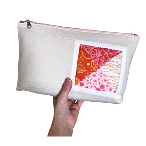 May include: Cream-colored canvas zippered pouch with a patchwork design. The patchwork features red, orange, and pink floral patterns. The zipper is silver with a pink trim. A hand is holding the pouch.