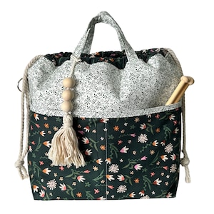 May include: A drawstring project bag featuring a dark green base with a multi-colored floral print and a white upper section with a black vine pattern. The bag includes two fabric handles, thick cream-colored rope drawstrings, and a wooden bead tassel.