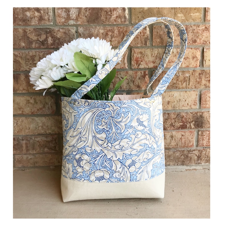May include: A blue and white floral tote bag with a cream coloured bottom. The bag has two handles and is filled with white flowers.