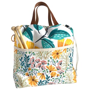Puede incluir: Una bolsa de lona blanca con estampado floral con flores amarillas, hojas verdes y detalles azules. La bolsa tiene un asa de cuero marr&oacute;n y un cierre de cord&oacute;n. Dos agujas de tejer de madera sobresalen de la parte superior de la bolsa.
