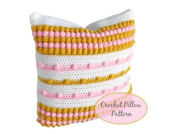 May include: A handmade crochet pillow with a white base, adorned with horizontal stripes of mustard yellow and pink bobbles and lines. The pillow has a textured appearance and the words "Crochet Pillow Pattern" are on the bottom right.