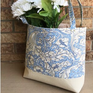 May include: A blue and white floral tote bag with a natural canvas base. The bag has two long straps and is filled with white flowers.