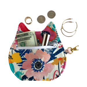 May include: A small, floral patterned cat-shaped zippered pouch with a pink and red ear, a white zipper, and a gold key chain. The pouch is open and contains a twenty dollar bill, a lipstick, and a gold ring.