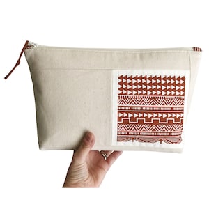 Pode incluir: Uma bolsa de lona creme com zíper e um patch retangular com um padrão geométrico em ferrugem e branco. A bolsa tem um puxador de zíper de couro marrom.