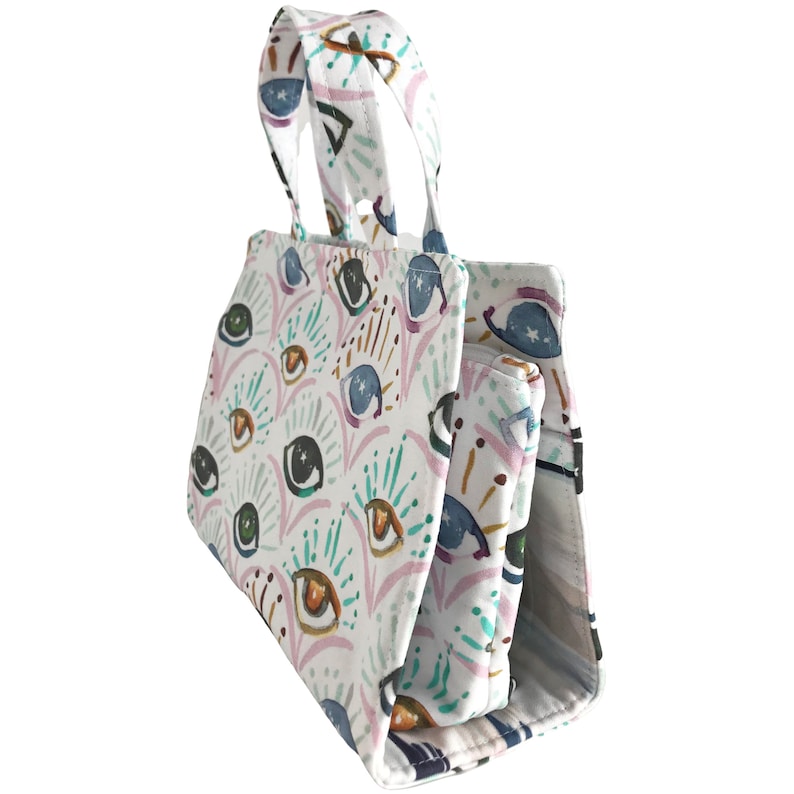 May include: A white tote bag with a colorful eye pattern. The bag has two handles and a zipper closure. The bag is made of a durable fabric and is perfect for carrying everyday essentials.