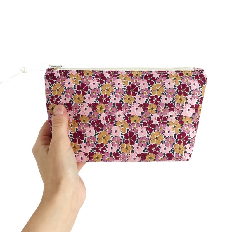 Puede incluir: Un neceser con estampado floral con cremallera y tirador blancos. La bolsa presenta un estampado de peque&ntilde;as flores rosas, burdeos y doradas sobre un fondo rosa claro. La bolsa se sostiene en una mano.