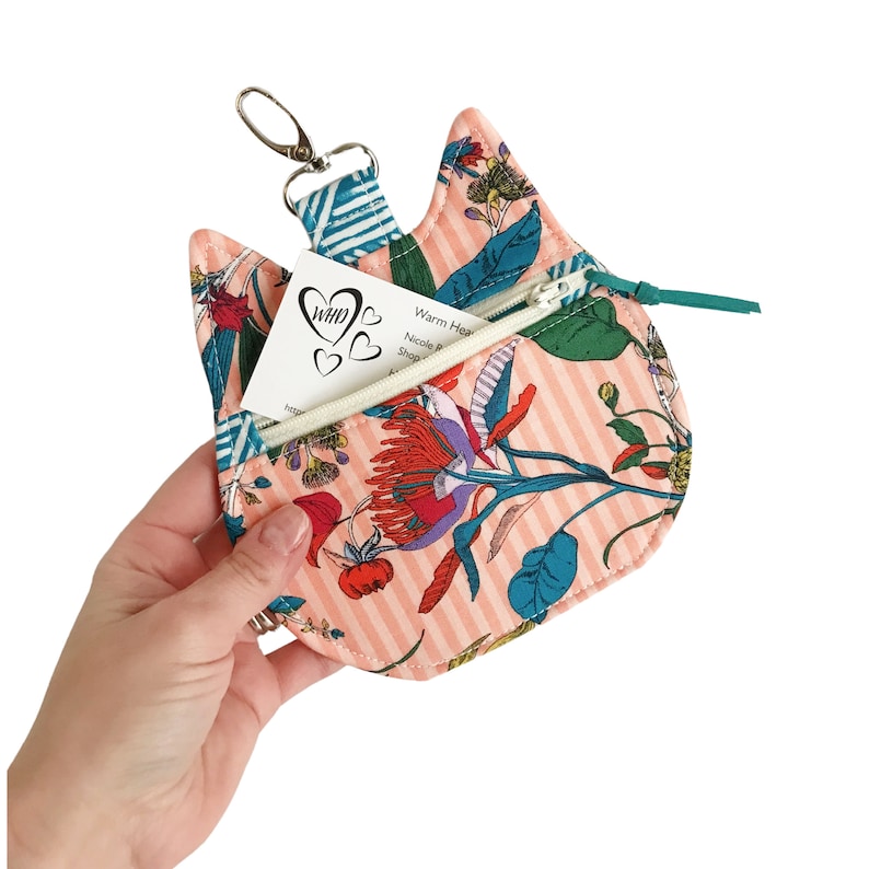 May include: A pink fabric cat-shaped keychain with a zippered pocket and a floral print. The keychain has a silver key ring and a white business card with the text "Warm Hearts Nicole P. Shop & Studio" inside the pocket.