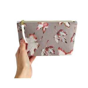 Puede incluir: Un neceser rectangular con fondo gris y estampado de hojas rojas, blancas y marrones. La bolsa tiene una cremallera dorada y una borla blanca. La bolsa est&aacute; sujeta por una mano.