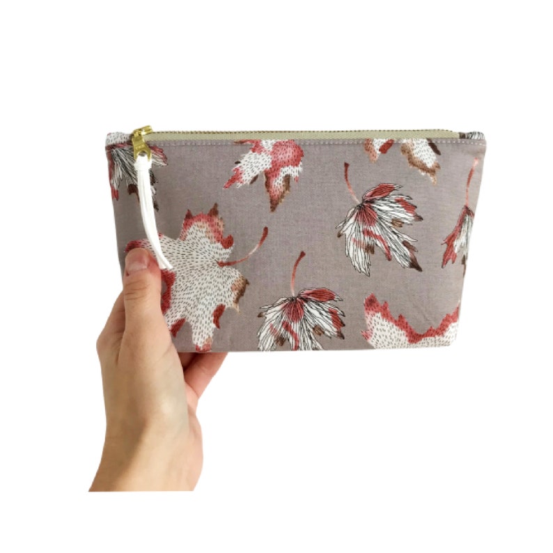 Puede incluir: Un neceser rectangular con fondo gris y estampado de hojas rojas, blancas y marrones. La bolsa tiene una cremallera dorada y una borla blanca. La bolsa est&aacute; sujeta por una mano.