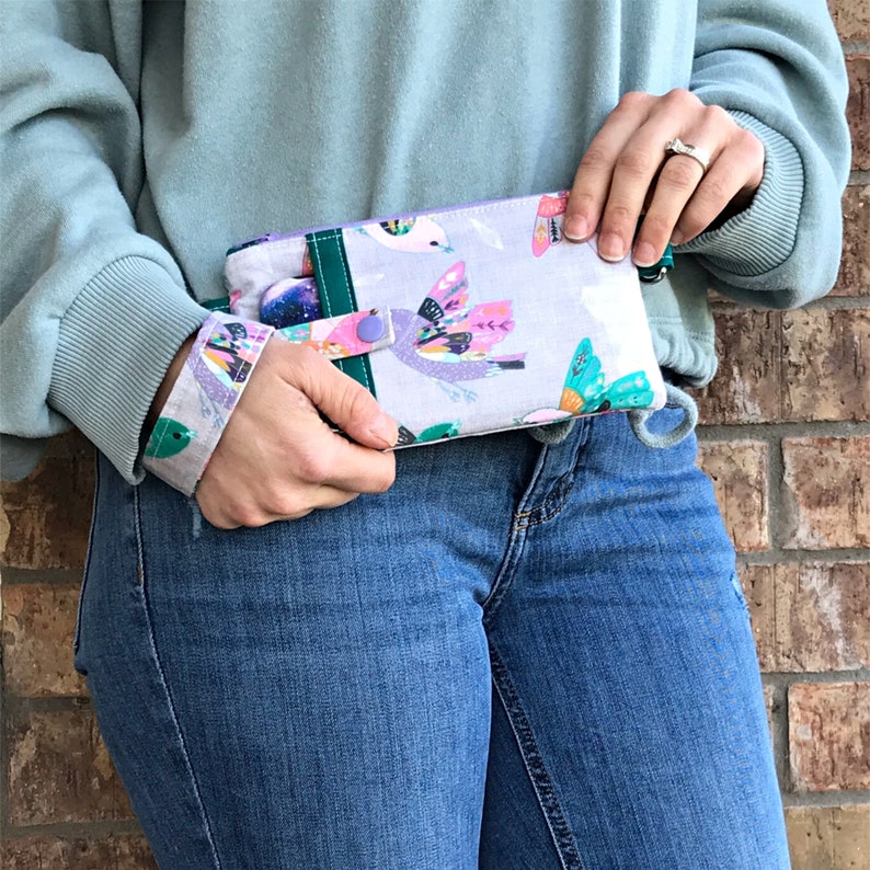 Puede incluir: Una mu&ntilde;equera de tela azul claro y verde con un bot&oacute;n morado y un estampado colorido de p&aacute;jaros y mariposas. La mu&ntilde;equera la lleva puesta una persona que lleva pantalones vaqueros azules.