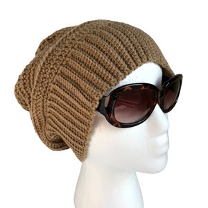May include: A brown crocheted beanie hat with a slouchy fit. The hat is worn on a white mannequin head with brown tortoise shell sunglasses.