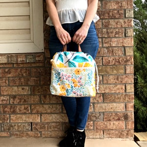 Puede incluir: Una bolsa de cord&oacute;n blanco con un patr&oacute;n floral con flores amarillas, hojas verdes y detalles blancos. La bolsa tiene un asa de cuero marr&oacute;n y un forro blanco.