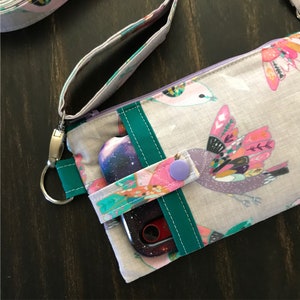 Puede incluir: Una funda de tel&eacute;fono de tela gris con cierre de cremallera y una correa verde azulada. La funda tiene un estampado de p&aacute;jaros coloridos con plumas. Un tel&eacute;fono est&aacute; dentro de la funda.