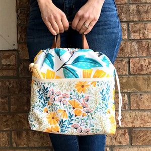 Puede incluir: Una bolsa de cord&oacute;n blanco con un patr&oacute;n floral en tonos amarillos, azules y verdes. La bolsa tiene un asa de cuero marr&oacute;n y un bolsillo separado en la parte delantera. Dos agujas de tejer de madera est&aacute;n dentro de la bolsa.