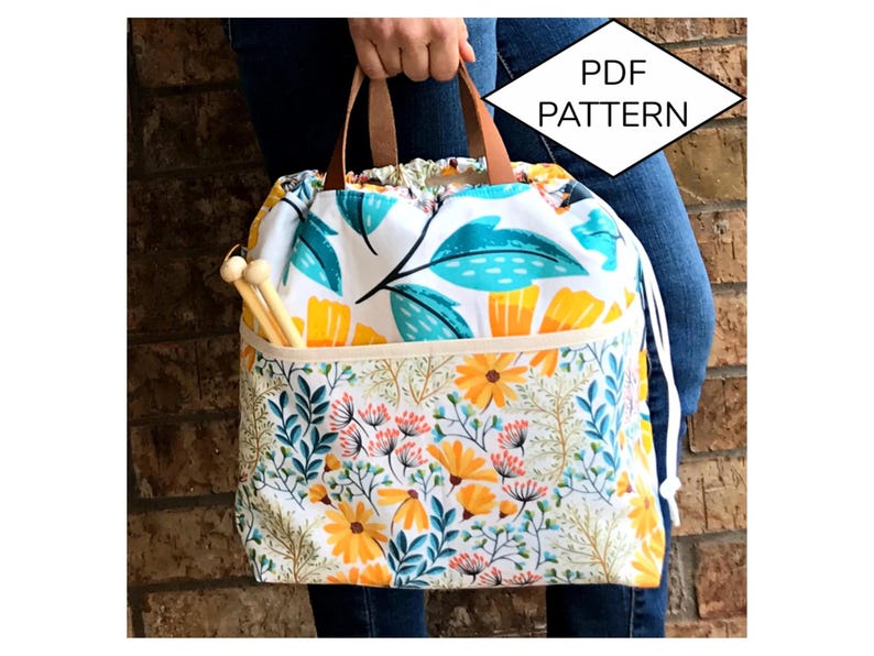 Puede incluir: Una bolsa de cord&oacute;n blanco con un estampado floral en tonos amarillos, azules y verdes. La bolsa tiene un asa de cuero marr&oacute;n y un cierre de cord&oacute;n blanco. La bolsa est&aacute; sentada sobre una superficie de ladrillo. El texto "PDF PATTERN" est&aacute; en forma de diamante sobre la bolsa.