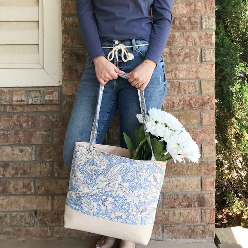 May include: A large, white canvas tote bag with a blue and white floral pattern. The bag is being held by a person wearing blue jeans and a blue shirt.