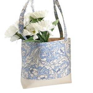May include: A blue and white floral tote bag with a cream coloured bottom. The bag has two handles and is filled with white flowers.