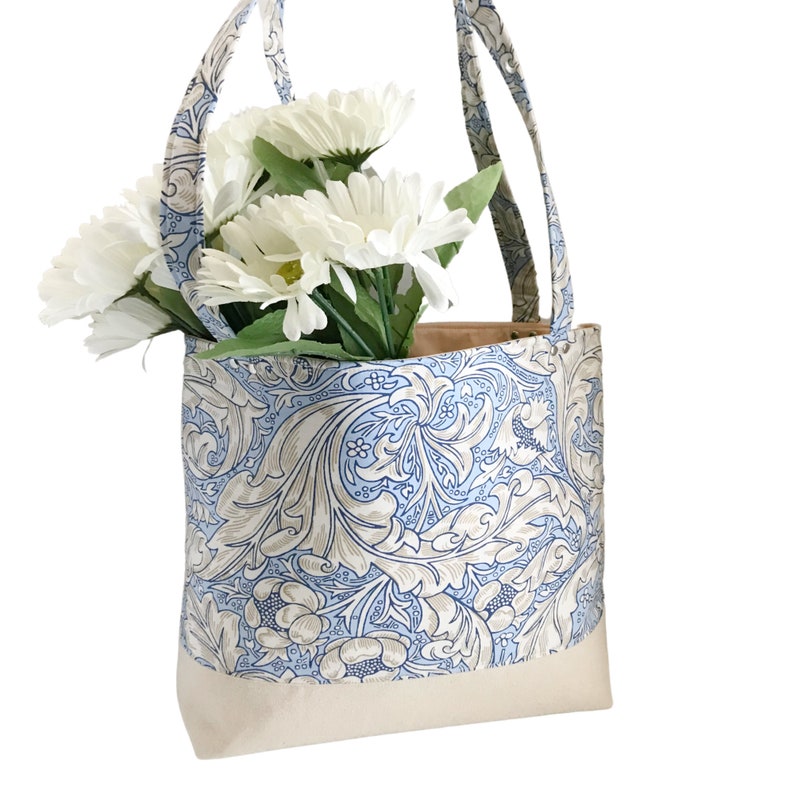 May include: A blue and white floral tote bag with a cream coloured bottom. The bag has two handles and is filled with white flowers.