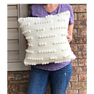 May include: A white crocheted pillow with a textured bobble stitch pattern. The pillow is being held by a person wearing a purple shirt and blue jeans.