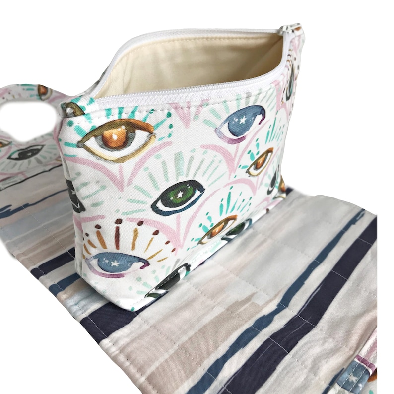 May include: A white zippered pouch with a colorful eye pattern. The pouch is open and the inside is lined with a cream-colored fabric. The pouch is sitting on top of a striped fabric.
