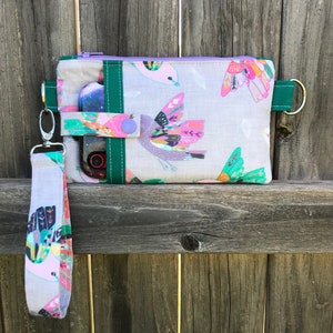 Puede incluir: Una bolsa de tela gris con cierre de cremallera y ribete verde. La bolsa presenta un estampado de p&aacute;jaros coloridos y una correa de mu&ntilde;eca desmontable. La bolsa se muestra en una valla de madera.