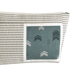 May include: A rectangular, gray and white striped fabric pouch with a teal pocket featuring white and black arrow patterns. The pocket is outlined with a white border. The pouch has a zipper closure.