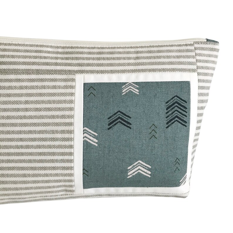 May include: A rectangular, gray and white striped fabric pouch with a teal pocket featuring white and black arrow patterns. The pocket is outlined with a white border. The pouch has a zipper closure.