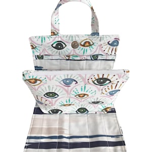 May include: A white canvas tote bag with a zippered pouch. The tote bag and pouch feature a colorful eye print with stars and swirls. The tote bag has two handles and a white and blue striped bottom.