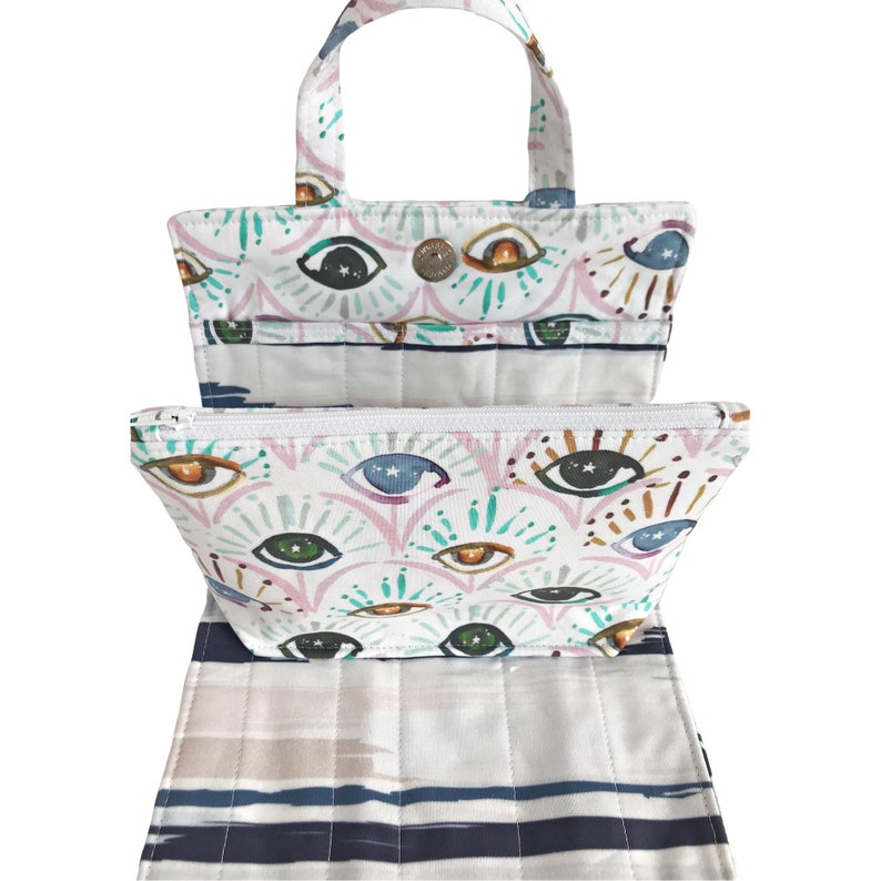 May include: A white canvas tote bag with a zippered pouch. The tote bag and pouch feature a colorful eye print with stars and swirls. The tote bag has two handles and a white and blue striped bottom.
