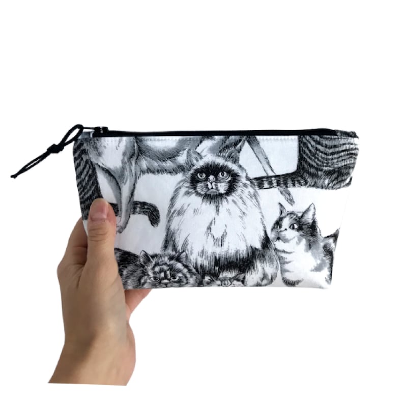 Puede incluir: Un neceser blanco con una ilustraci&oacute;n en blanco y negro de varios gatos. La bolsa tiene una cremallera negra y un cord&oacute;n negro. Los gatos est&aacute;n representados en varias poses, con pelaje y expresiones detalladas.