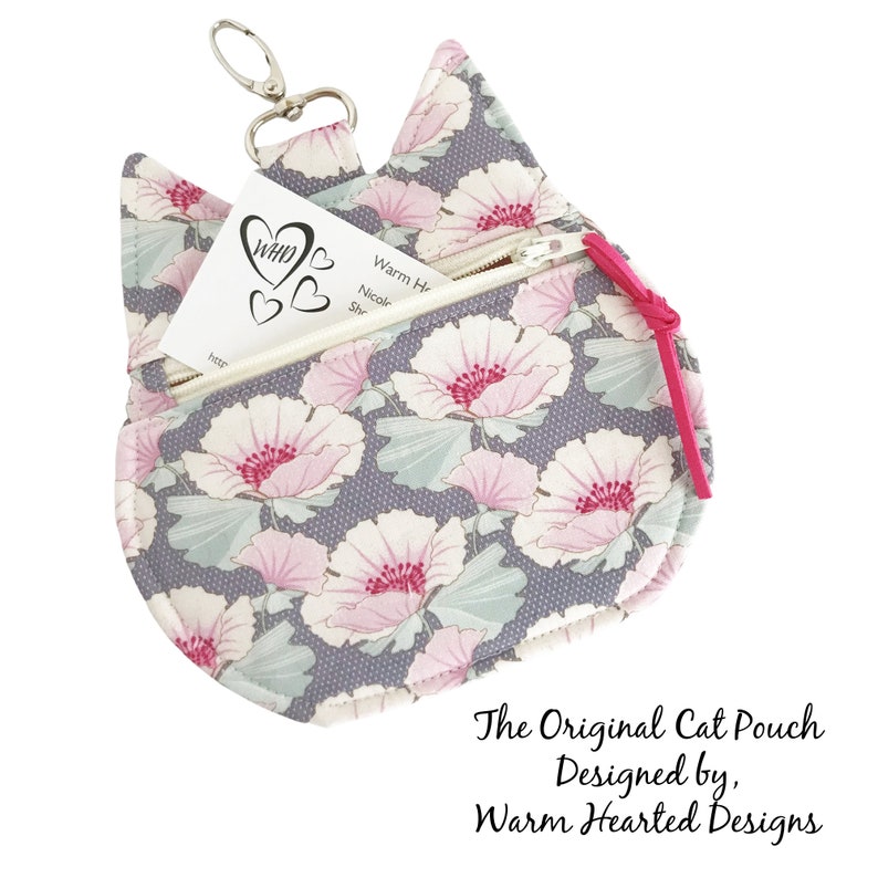 May include: A gray cat-shaped pouch with a pink floral pattern and a pink zipper. The pouch has a key ring and a small card inside that says "WHD" with hearts. The text "The Original Cat Pouch Designed by, Warm Hearted Designs" is below the pouch.