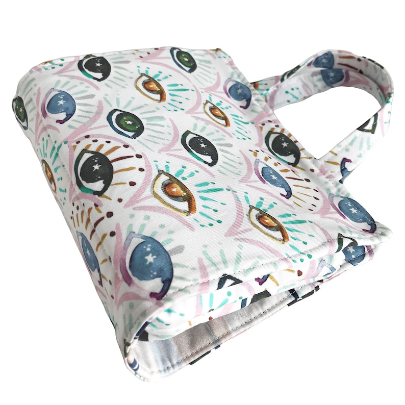 May include: A white tote bag with a colorful eye pattern. The eyes are surrounded by a variety of colors, including pink, blue, green, and brown. The bag has a single handle.