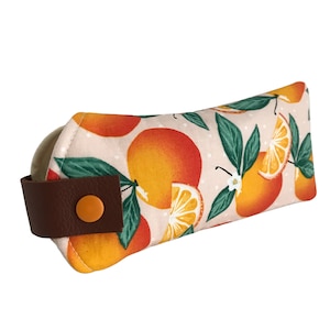 Pode incluir: Uma m&aacute;scara para os olhos de tecido rosa claro com uma al&ccedil;a de couro marrom e um bot&atilde;o laranja. O tecido tem um padr&atilde;o de fatias de laranja e folhas sobre um fundo branco.