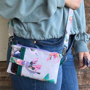 Puede incluir: Una peque&ntilde;a bolsa de tela gris y blanca con una cremallera morada y una correa verde y blanca. La bolsa tiene un estampado floral con p&aacute;jaros y mariposas. Se puede ver un tel&eacute;fono dentro de la bolsa.