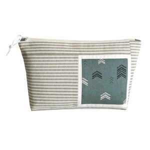 May include: A rectangular cosmetic bag with a gray and white striped pattern. A teal square with white and black arrow designs is on the front. The bag has a white zipper and a white pull.