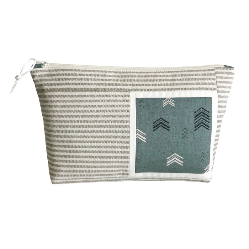 May include: A rectangular cosmetic bag with a gray and white striped pattern. A teal square with white and black arrow designs is on the front. The bag has a white zipper and a white pull.
